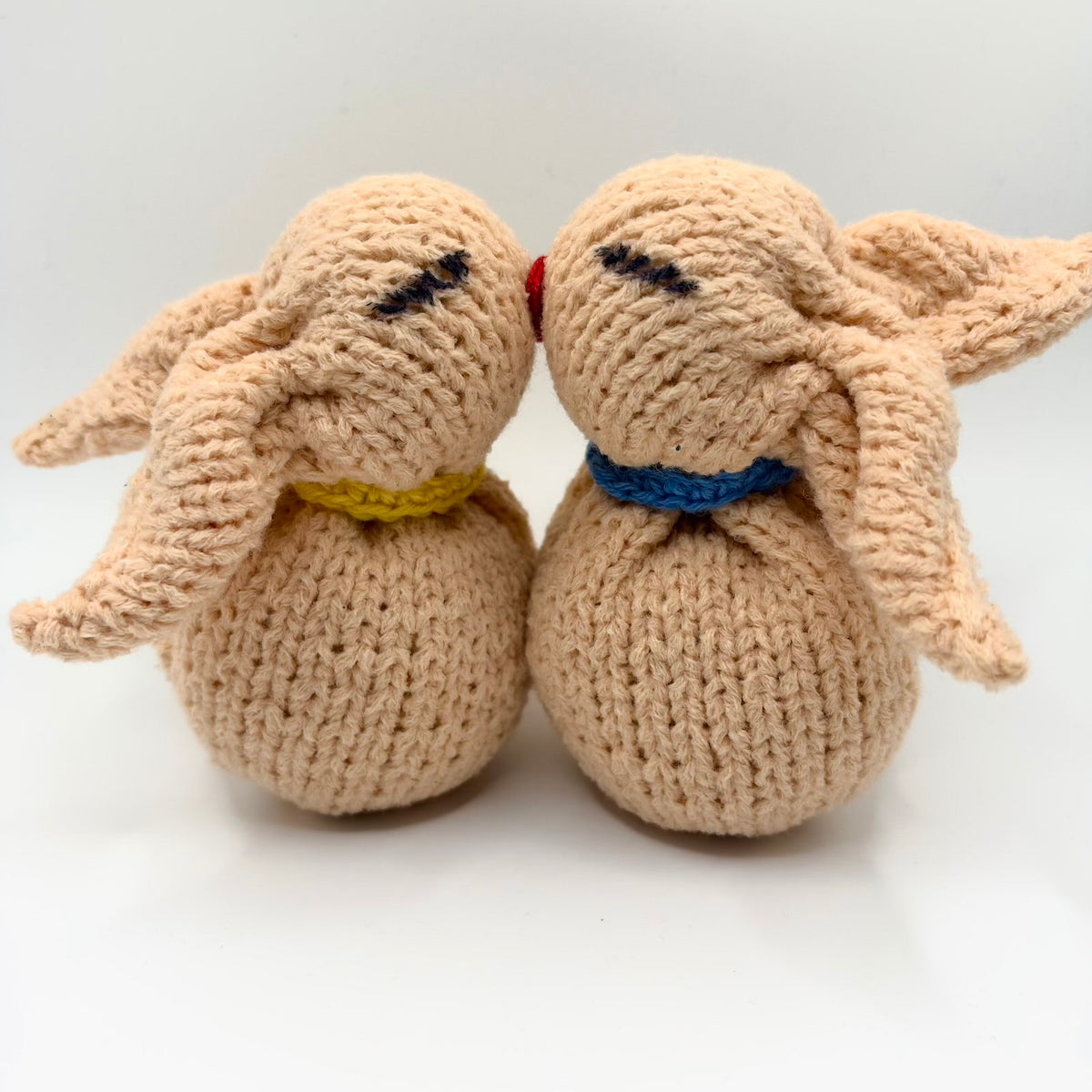 Two peachy-beige knitted bunny plushies nestled together, with long floppy ears, rounded bodies and closed embroidered eyes. One wears a yellow collar and the other a blue collar.