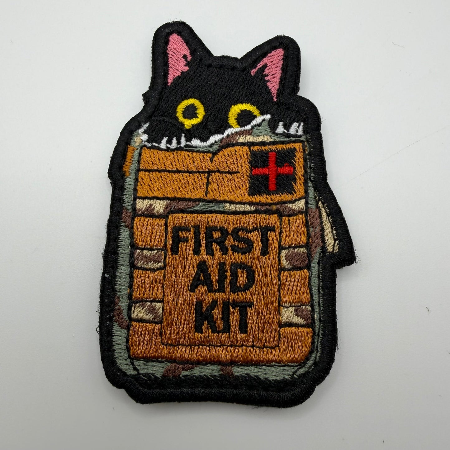 Embroidered patch of a cat peeking out from a first aid kit on a white background