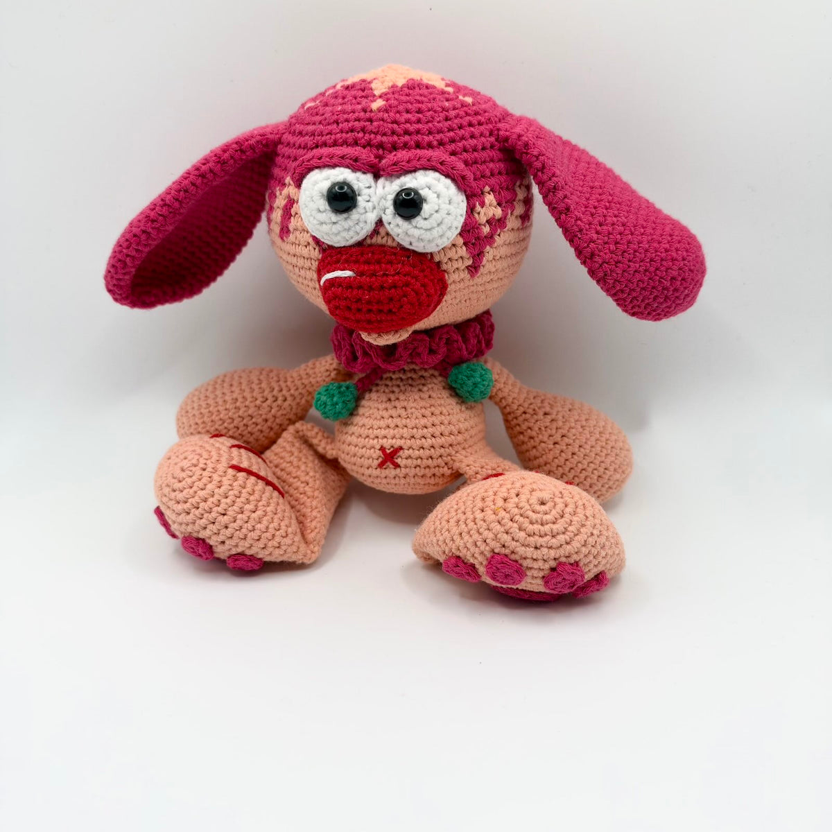 Peach-coloured crochet character with long raspberry-pink floppy ears, oversized white eyes with black pupils and a large red nose. It wears a pink ruffled collar with two green bobbles and has large paws with pink toes and red stitched details.