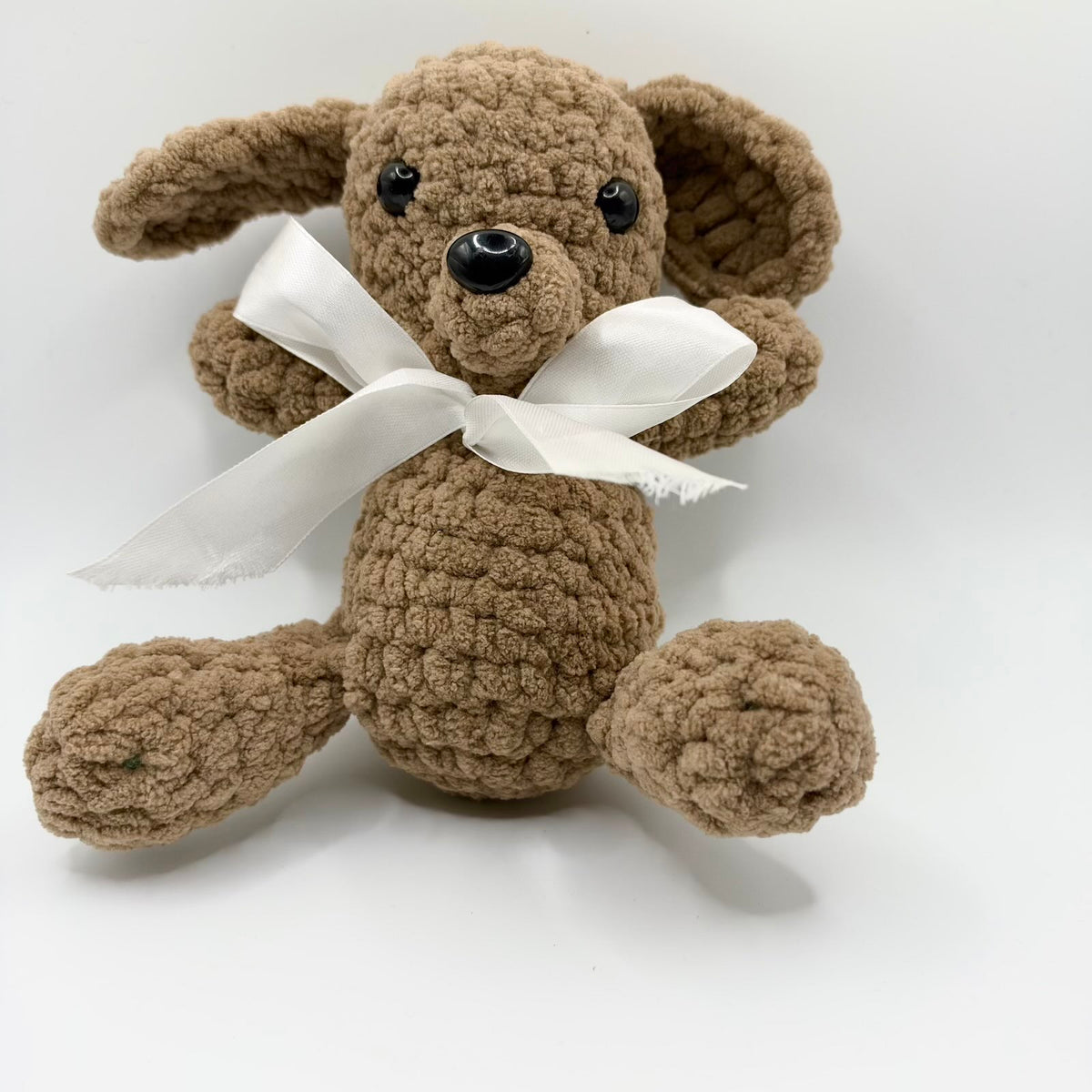 Brown crochet puppy plush toy sitting against a plain white background, with large floppy ears, shiny black eyes and nose, a rounded muzzle and outstretched legs with broad paws. A large white ribbon is tied in a bow around its neck, and its short arms extend to either side.