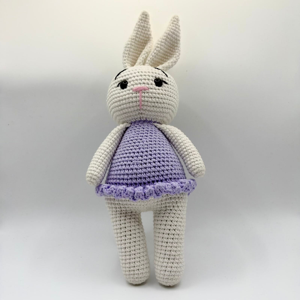 Cream-coloured crochet bunny with long upright ears, shiny black eyes, embroidered black eyelashes and eyebrows, and a pink stitched nose and mouth. It wears a lilac crochet dress with a ruffled hem and has long cream arms and legs, photographed from the front against a plain light background.