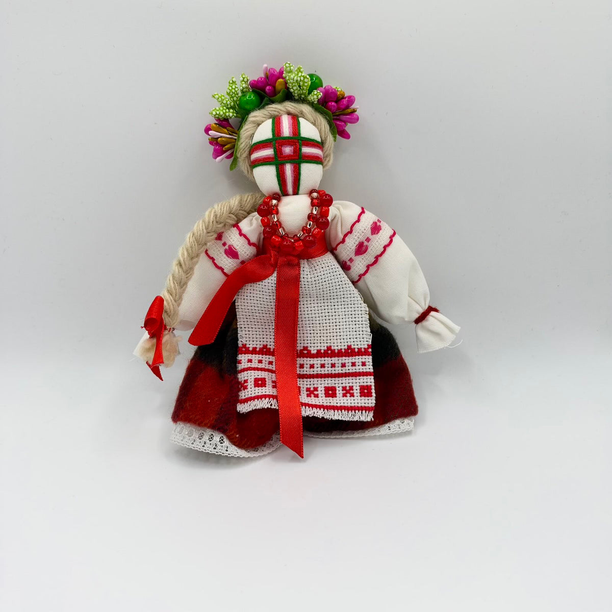 Ukrainian Motanka cloth doll with a multicoloured thread cross covering her face, a long fair yarn plait and a pink-and-green floral wreath. She wears red beads, a white blouse with red stitching, a red ribbon sash, an embroidered white apron and a red-and-black skirt trimmed with white lace.