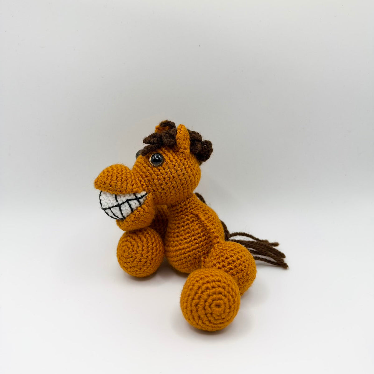 Golden-orange crochet horse sitting at an angle against a white background, with a long muzzle and an oversized white grin divided into teeth with black stitching. It has shiny eyes, upright ears, a curly dark brown mane, a loose brown yarn tail and chunky rounded hooves.