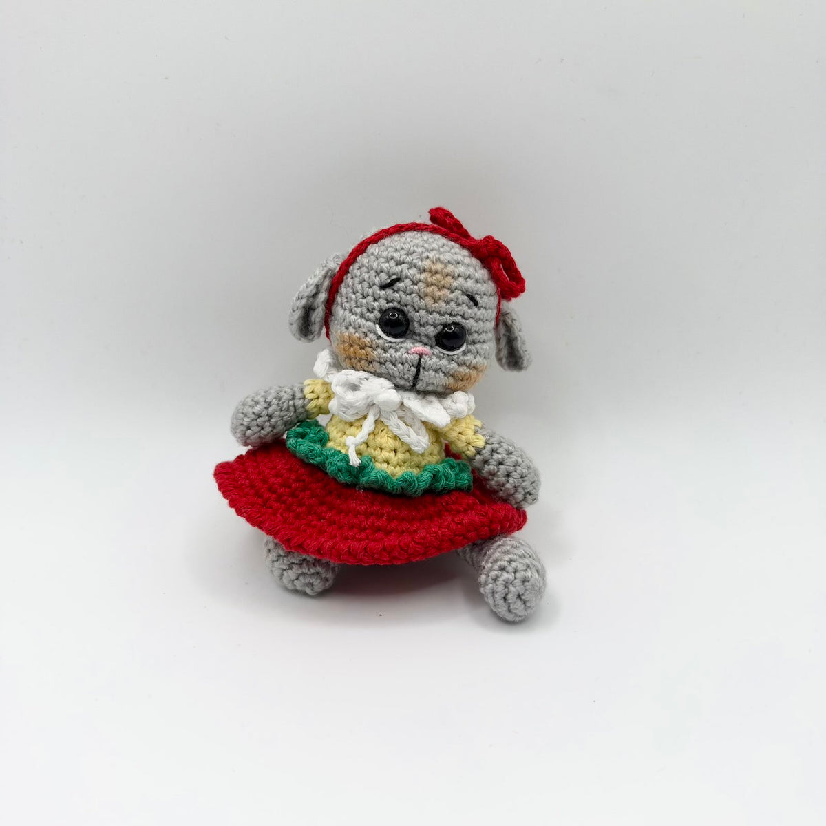 Grey crochet puppy with floppy ears, tan patches on its face, large shiny black eyes, embroidered eyebrows and a pink nose. It wears a red bow headband, pale yellow top with green ruffled trim, white frilly collar and flared red skirt, sitting with its head tilted against a white background.