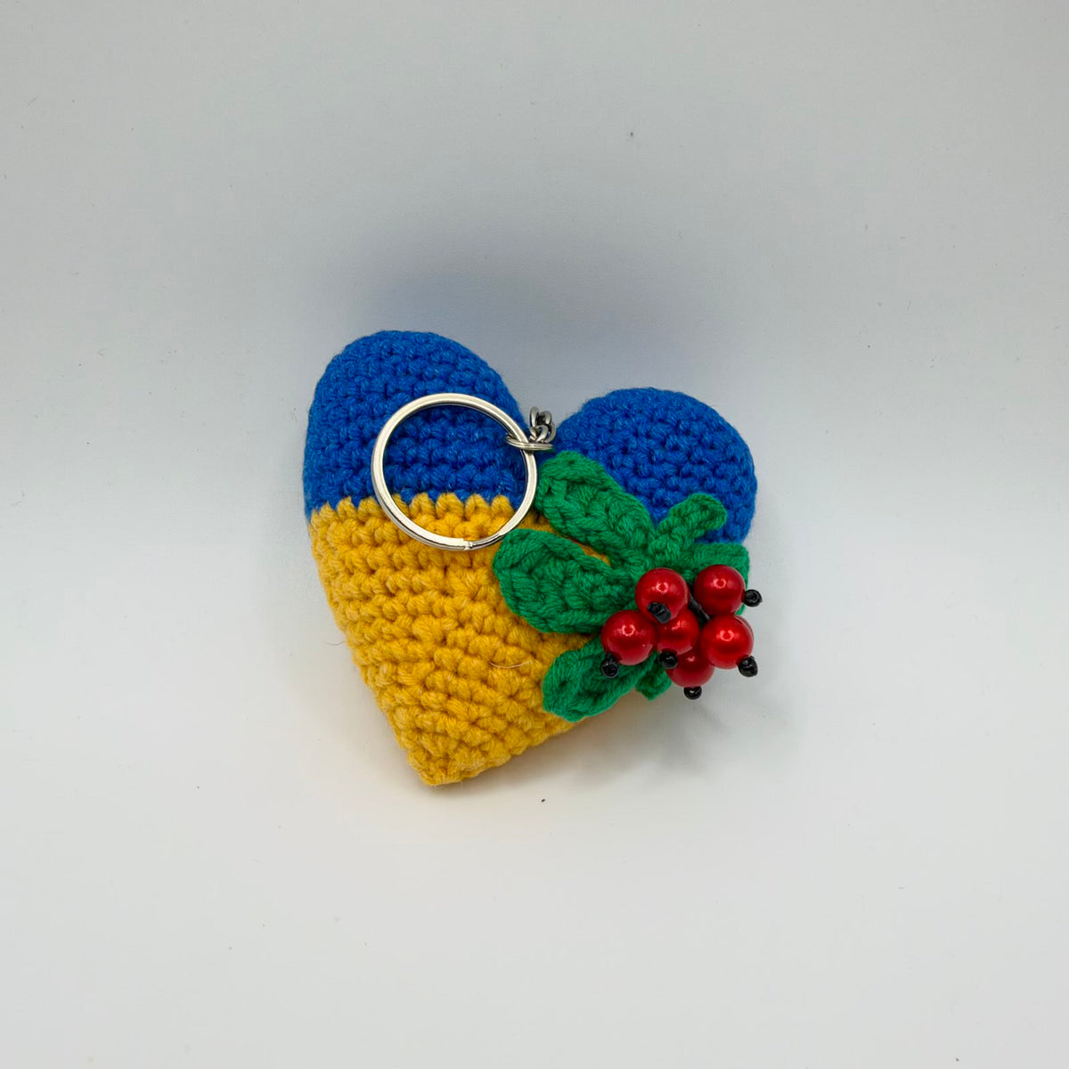 Crochet heart keyring with a blue upper half and yellow lower half, decorated on one side with green crochet leaves and a cluster of glossy red berries with black tips. A silver-coloured split keyring is attached at the top. Photographed against a plain light background.