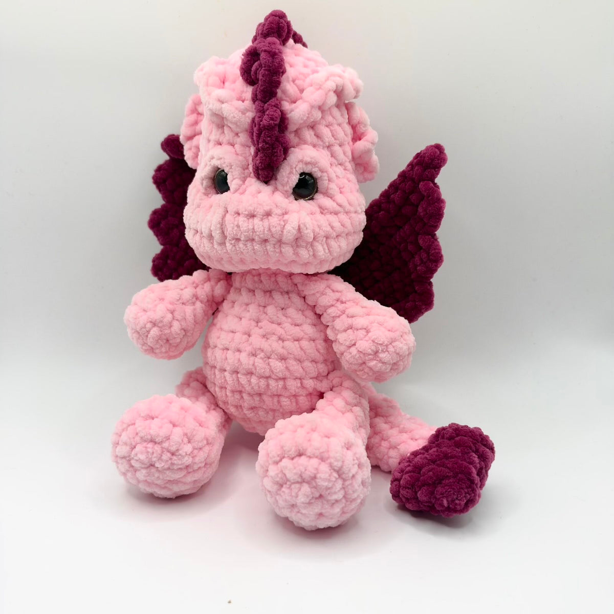 ink crochet dragon plushie sitting against a white background, with deep purple wings, a purple crest along its head and a purple tip on its long pink tail. It has shiny dark eyes, a broad rounded muzzle, small arms and large rounded feet, with chunky plush-textured stitches throughout.