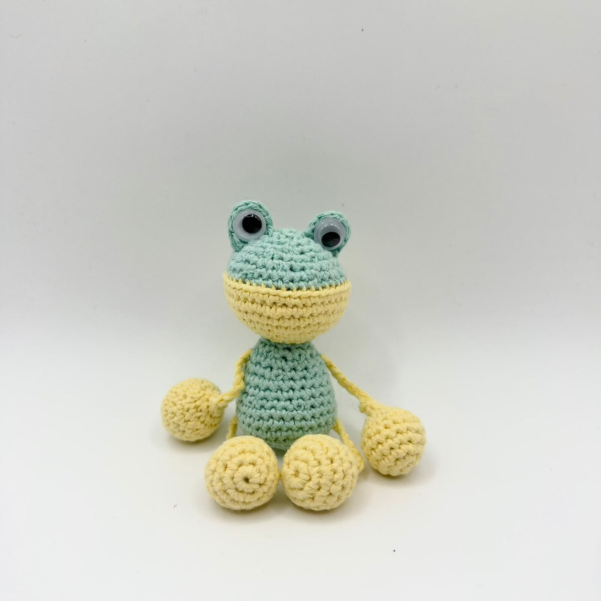 Small mint-green crochet frog with raised googly eyes and a pale yellow lower face. Thin yellow yarn limbs connect its body to large rounded yellow hands and feet. The frog is seated facing the camera against a plain light background.