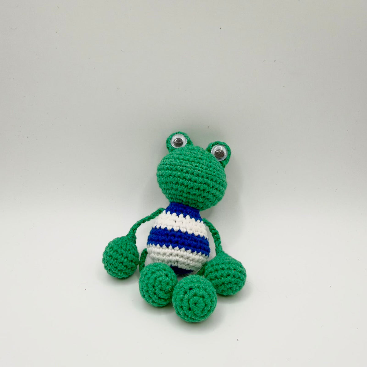 Green crochet frog with raised googly eyes and a rounded body patterned with broad blue-and-white horizontal stripes. Thin green yarn limbs connect to large rounded green hands and feet. The frog sits with its head tilted slightly, photographed against a plain light background.