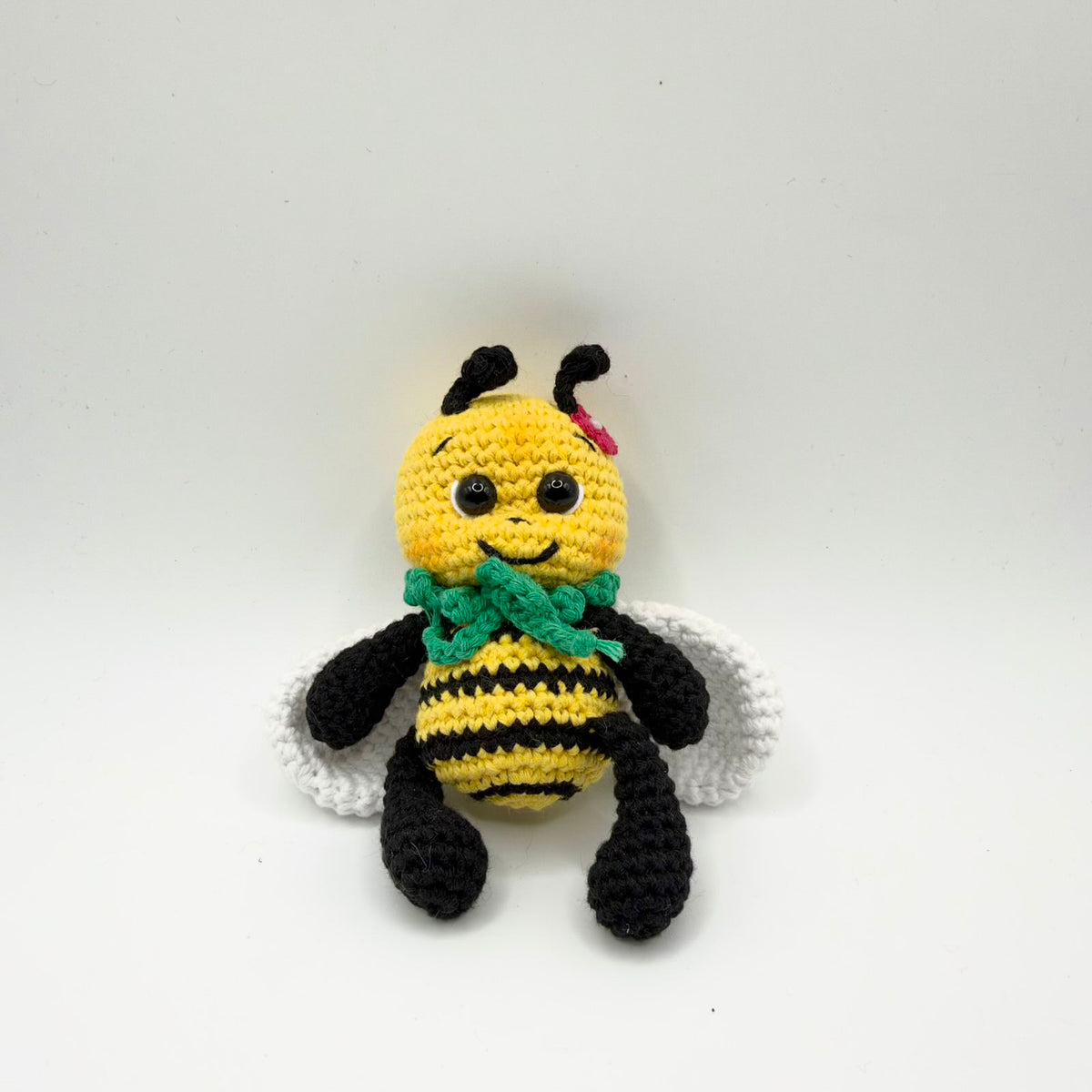 Crochet bee with a yellow head, curled black antennae, shiny dark eyes and an embroidered smile. It has a small pink flower beside one antenna, a green ruffled collar, a yellow-and-black striped body, white wings, black arms and long black legs. Seated against a plain light background.