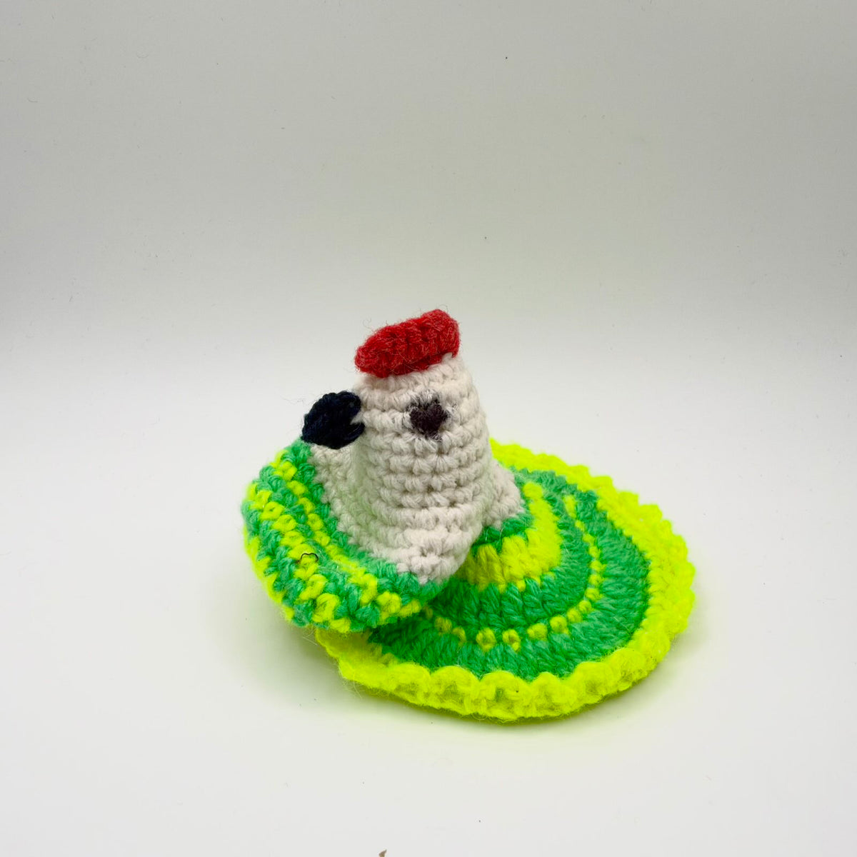 Crochet hen-shaped egg warmer with a cream head and neck, red comb, dark beak and dark stitched eye. Its green body and patterned wings are decorated with vivid yellow stitches, with a broad fan-shaped tail and bright yellow scalloped edging. Shown against a plain light background.