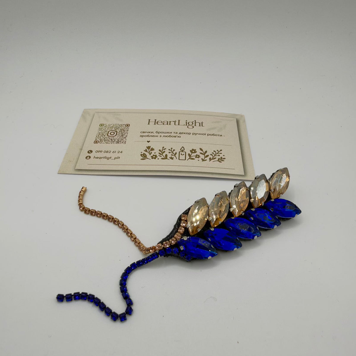 Wheat-shaped brooch with two rows of elongated faceted stones in royal blue and pale champagne, with matching strands of small decorative stones extending from the base. Shown on a light surface beside a HeartLight maker’s card.