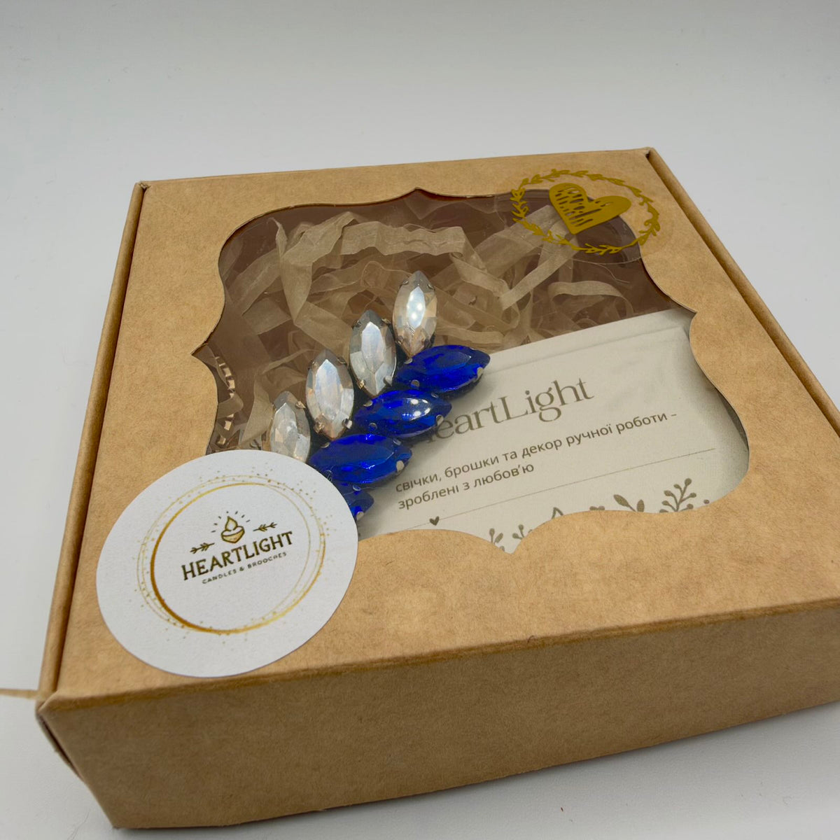 Blue and champagne-coloured wheat brooch nestled in shredded paper inside a brown presentation box with a decorative clear window. A HeartLight maker’s card is visible inside, with a round HeartLight label and gold heart decoration on the lid.