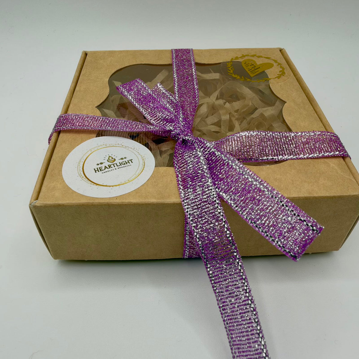 Brown HeartLight presentation box with a decorative clear window, tied with a shimmering purple ribbon edged in silver. The lid carries a round white HeartLight label and a gold heart decoration, with shredded paper packaging visible through the window.