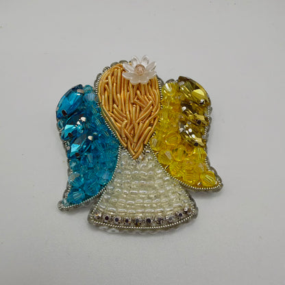 Angel-shaped brooch with turquoise-blue beads and faceted stones on the left wing and yellow beads and stones on the right. Gold-coloured textured detailing forms the centre beneath a translucent flower. An ivory beaded skirt, sparkling lower border and silver-coloured edging complete the design.