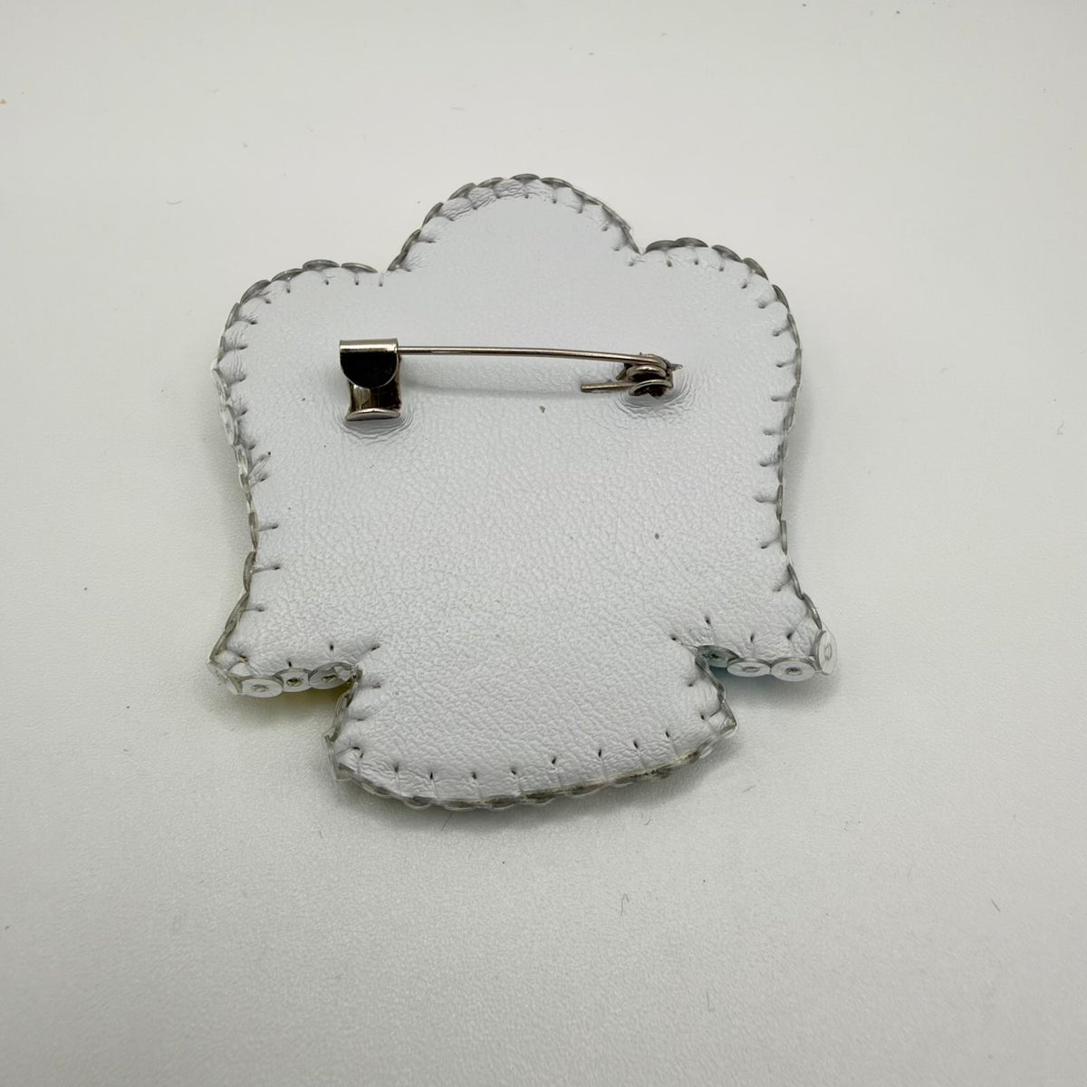 Reverse of the angel-shaped brooch showing a white textured backing with visible stitching around the outline. A horizontal silver-coloured brooch pin and clasp sit across the upper section, with translucent edging beads visible around the perimeter.