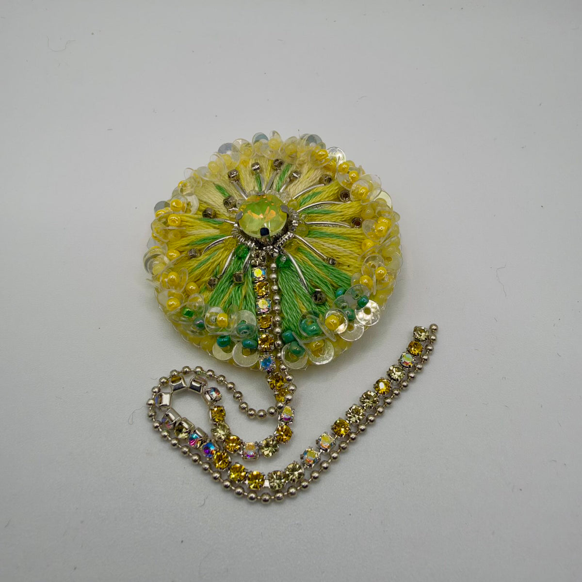 Round floral brooch with yellow, ivory and green threads radiating from a yellow-green faceted centre stone. Translucent sequins, small beads and silver-coloured details decorate the bloom. A strand of yellow and iridescent stones and a silver-coloured ball chain trail below it on a light background.