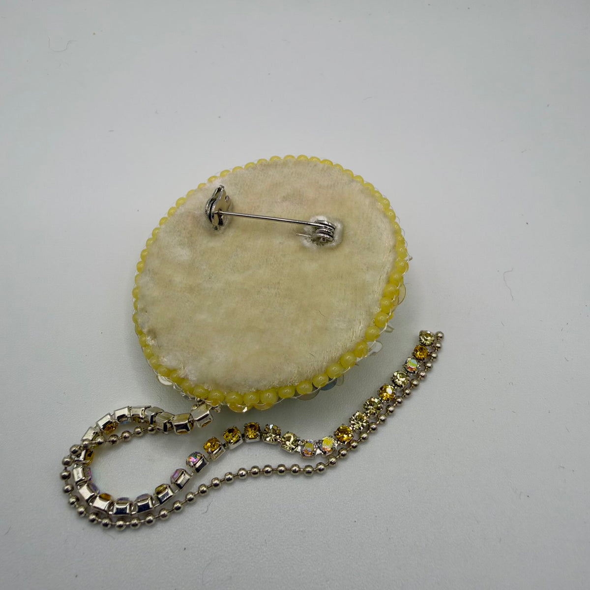 Reverse of the round brooch showing a cream felt-like backing and a horizontal silver-coloured pin fastening near the top. Yellow beads and translucent sequins edge the perimeter. Two decorative strands, one set with coloured stones and one a silver-coloured ball chain, extend below.
