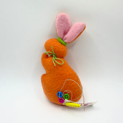 Orange felt-like bunny in a seated side profile, with tall pink-lined ears gathered at the base with green trim. A green cord bow is tied around its neck. Blue, green and pink buttons decorate its lower body beside a yellow handmade tag and a paper greeting.