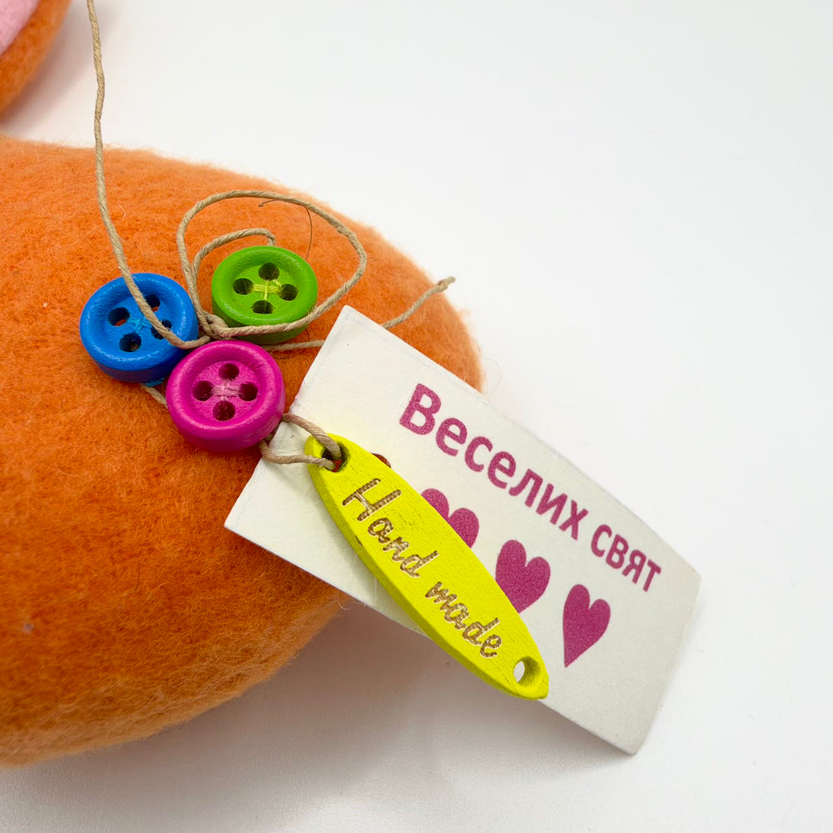 Close-up of blue, green and pink buttons attached to the orange bunny, with decorative twine and a yellow tag reading “Hand made”. Beside them, a white paper tag with pink hearts reads “Веселих свят”, Ukrainian for “Happy holidays”.