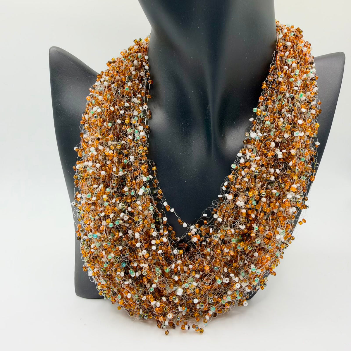Full multistrand beaded necklace displayed on a black jewellery bust. Numerous fine translucent looped strands form a broad cascading shape, scattered with small amber-orange, brown, milky-white and iridescent green-toned beads.