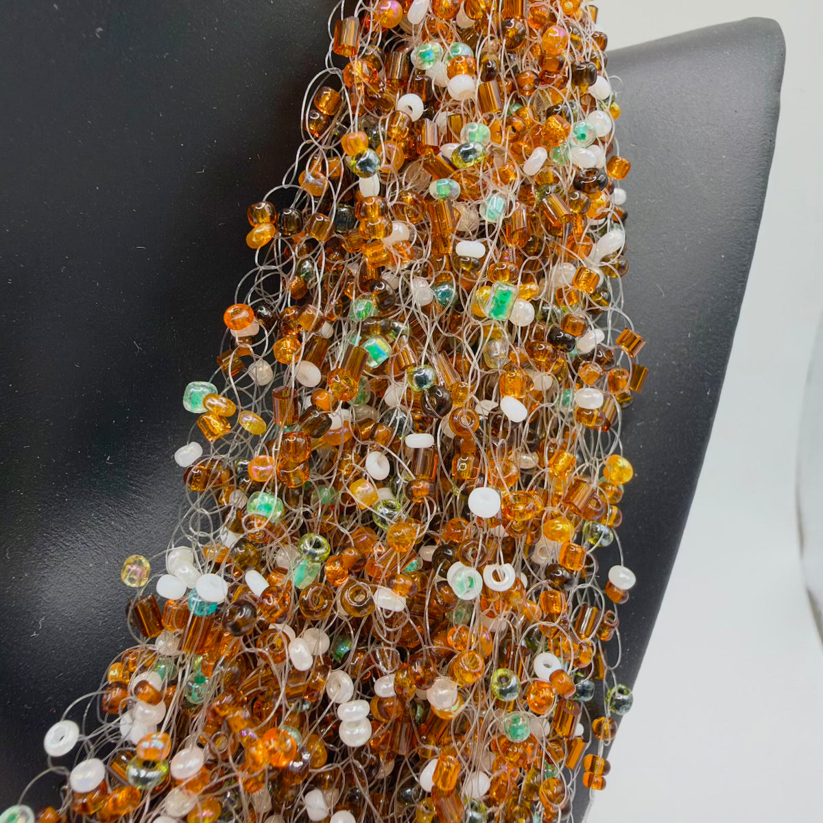 Close-up of the necklace’s layered beadwork against a black display bust, showing fine translucent loops threaded with small round and tubular beads in honey-orange, brown, white and shimmering pale green tones.