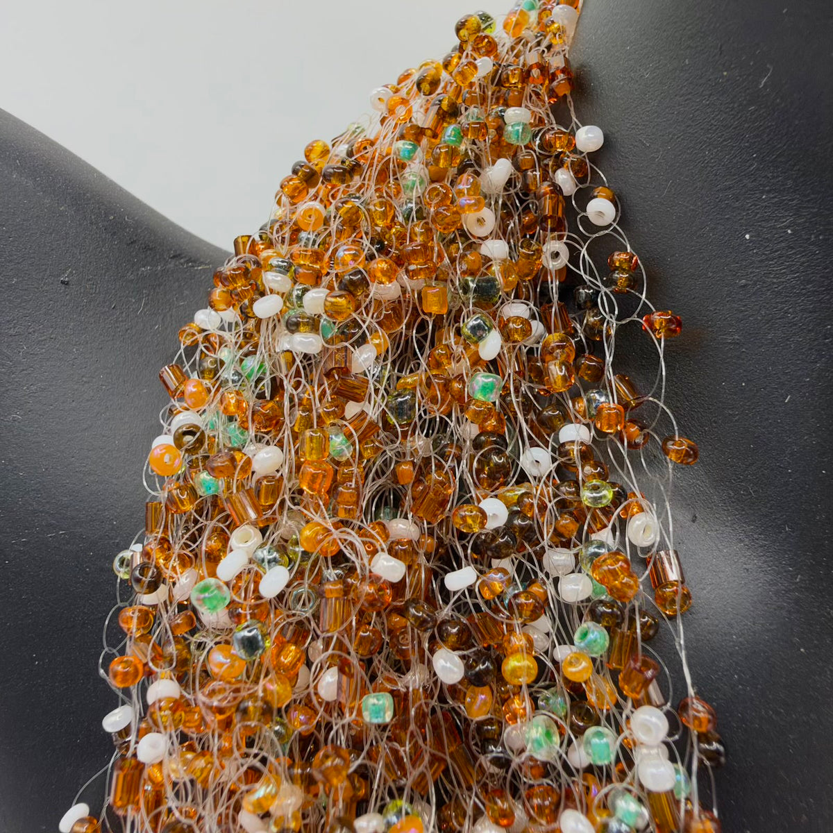 Angled detail of the necklace draped over a black jewellery bust, showing its airy network of translucent strands and densely scattered amber-coloured, white, brown and green-iridescent beads in varied shapes.