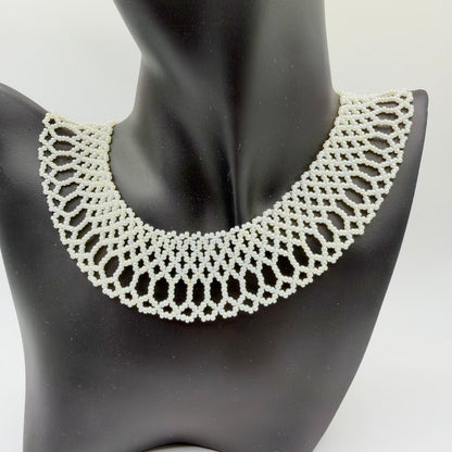 Ivory-coloured beaded collar necklace displayed on a black jewellery bust. Small lustrous beads form a broad, symmetrical openwork design with interlocking geometric shapes, elongated loops and a softly scalloped lower edge.