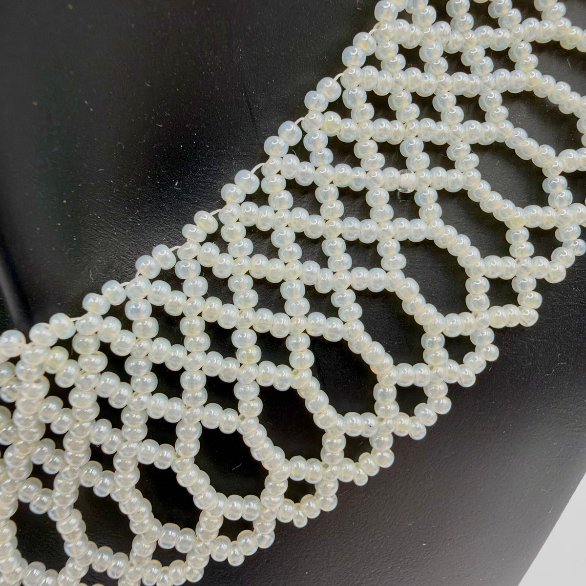 Close-up of the necklace’s ivory-coloured beads against a black display bust, showing their pearly sheen and intricate lace-like construction. Linked geometric openings lead into elongated beaded loops with small scallops along the outer edge.