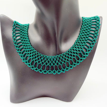 Emerald-green beaded collar necklace displayed on a black jewellery bust. Small lustrous beads form a broad curved band of interlocking geometric openwork, with elongated loops and a scalloped outer edge. The green beads show teal highlights in the light.