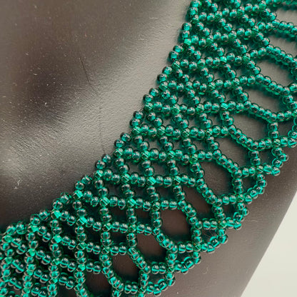 Close-up of translucent emerald-green beads forming the necklace’s lace-like pattern against a black display bust. Rows of linked geometric openings lead into long rounded loops and small scallops along the lower edge.