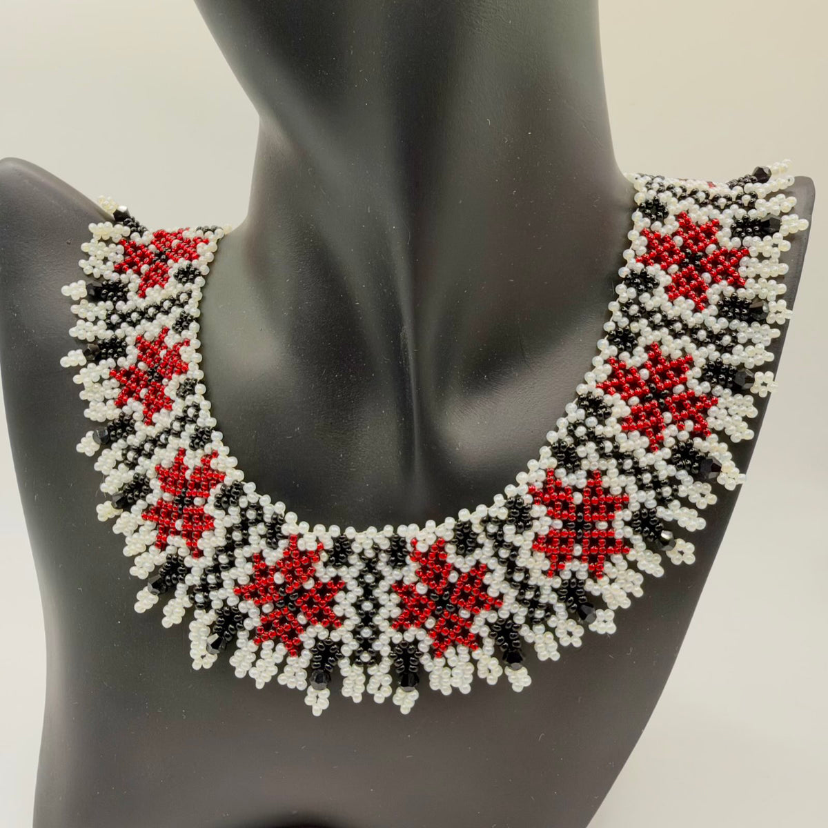 Broad beaded collar necklace displayed on a black jewellery bust. Repeating red floral motifs sit within an ivory-and-black geometric pattern. The curved lower edge is finished with small ivory beaded loops and dangling faceted black beads.