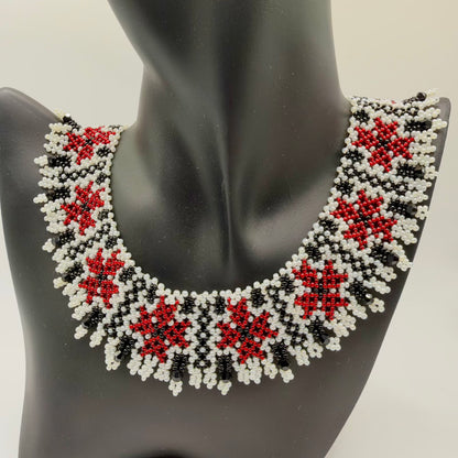 Broad beaded collar necklace displayed on a black jewellery bust. Repeating red floral motifs sit within an ivory-and-black geometric pattern. The curved lower edge is finished with small ivory beaded loops and dangling faceted black beads.