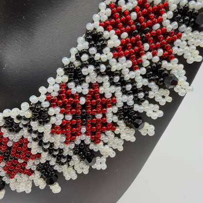 Close-up of the necklace’s red flower motifs, glossy black geometric details and pearly ivory-coloured beads. Small looped bead fringes and larger faceted black drops decorate the lower edge, shown against a black display bust.