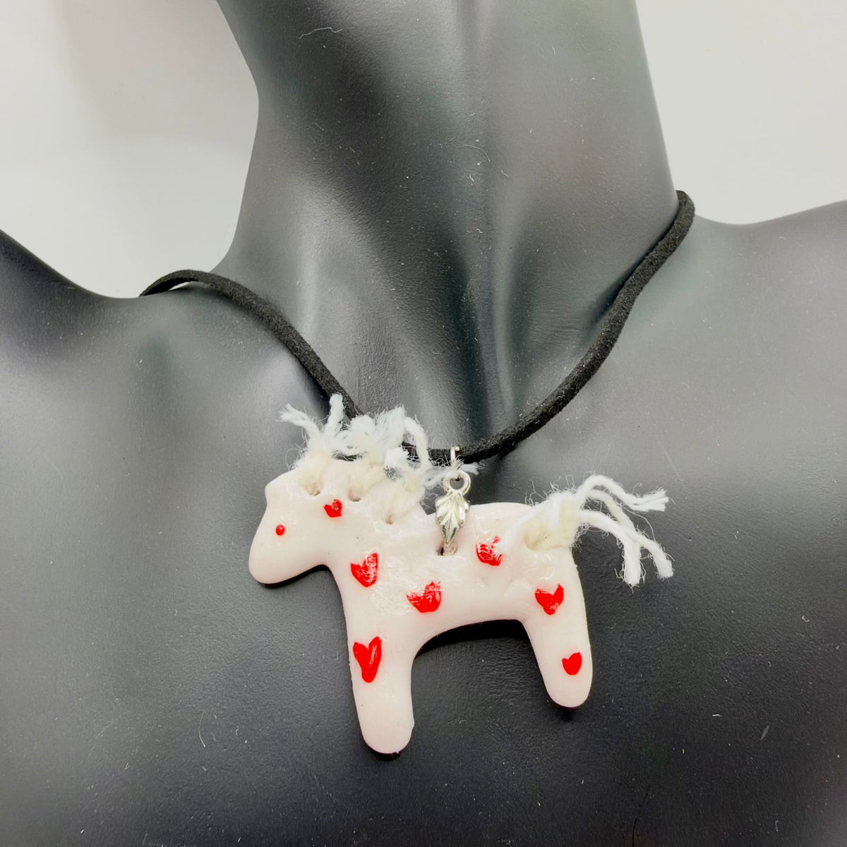 Pale blush-white horse-shaped pendant decorated with small red hearts, a red eye and a white yarn mane and tail. It hangs from a black cord through a silver-coloured bail, displayed against a black jewellery bust.