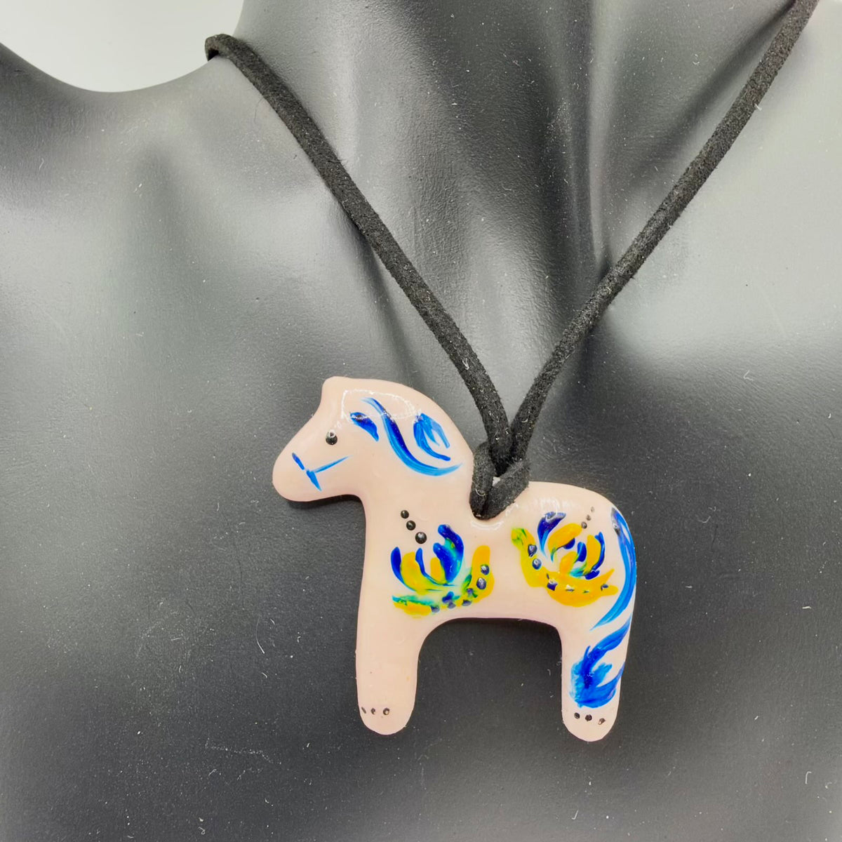 Glossy pale blush horse-shaped pendant on a black cord, displayed against a black jewellery bust. The horse is decorated with blue-and-yellow floral brushwork, a flowing blue painted mane and tail, a black eye and small black dotted accents on its neck, back and legs.