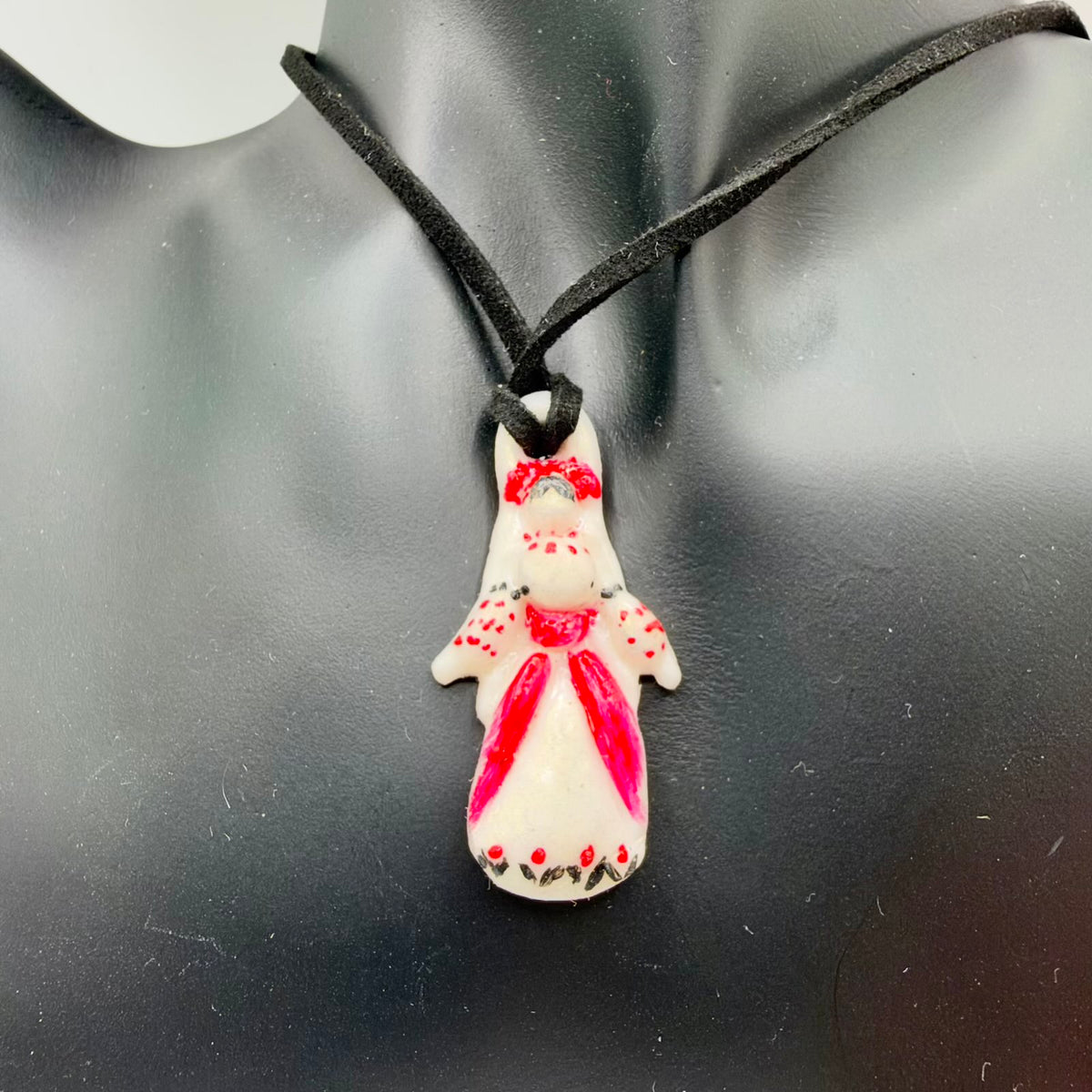 Ivory-coloured pendant shaped like a woman in Ukrainian folk-inspired dress, hanging from a black cord on a black display bust. The glossy figure has a red floral headdress, red skirt panels, dotted sleeves and red flowers with dark leaves around the hem.