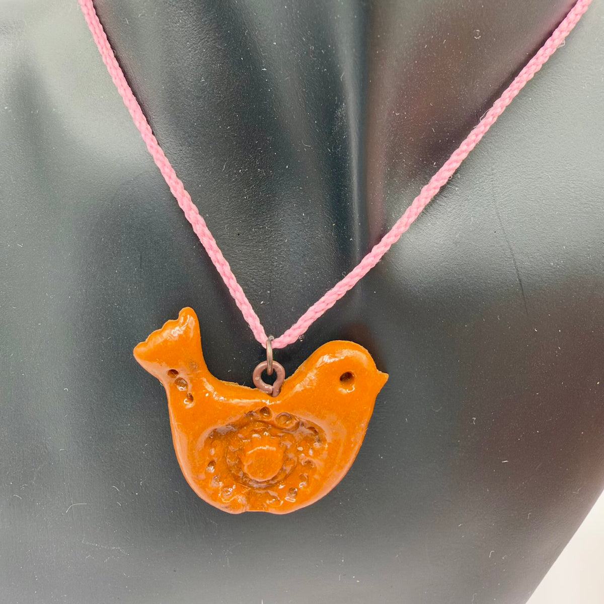 Glossy orange bird-shaped pendant hanging from a pink braided cord on a black jewellery bust. The bird faces right, with a small pointed beak, indented eye, raised tail and a textured decorative wing pattern. A metal ring connects the pendant to the cord.