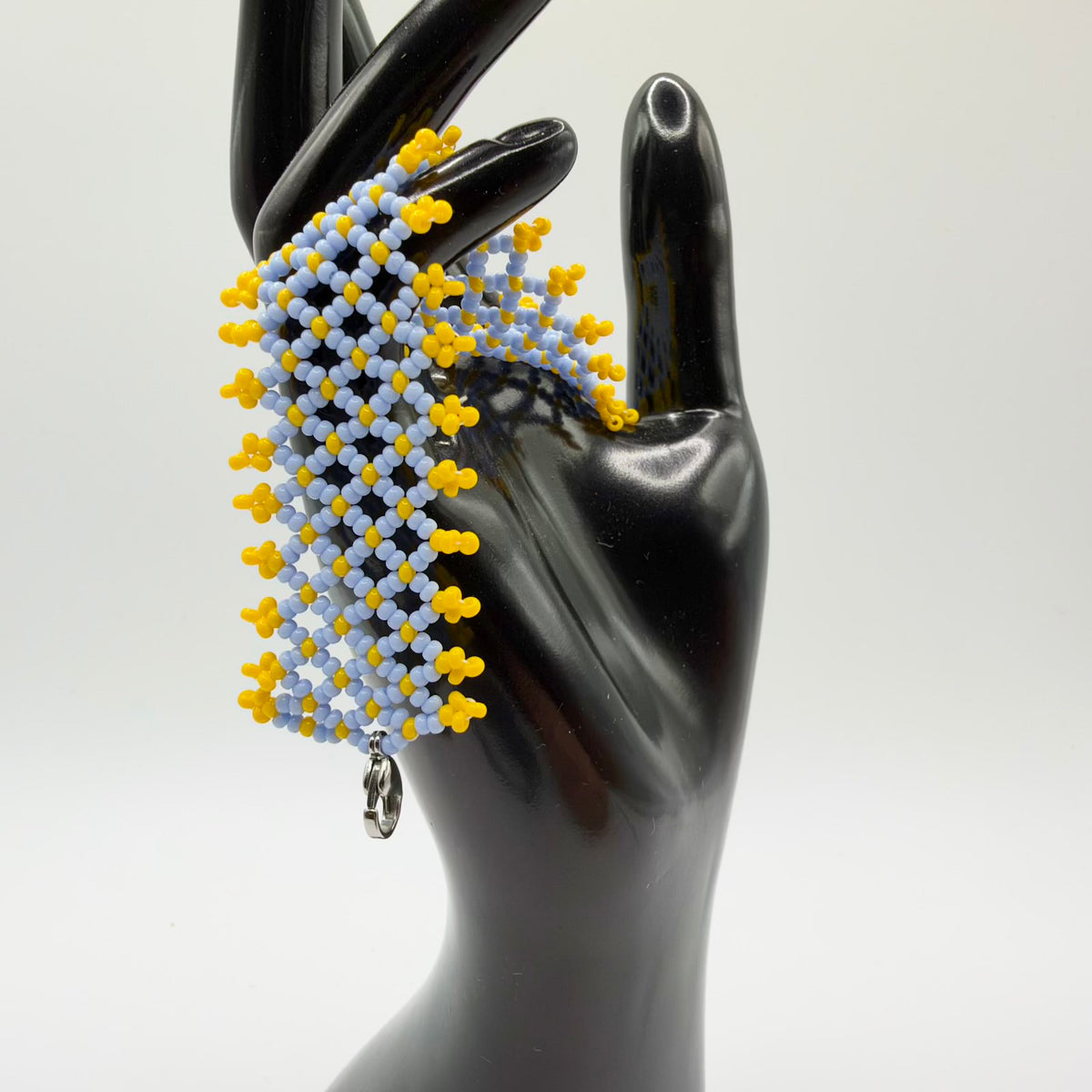 Wide beaded bracelet draped over a black hand-shaped display. Pale blue beads form an open diamond lattice with yellow beads at the intersections and small yellow clusters along both edges. A silver-coloured clasp hangs from the lower end.