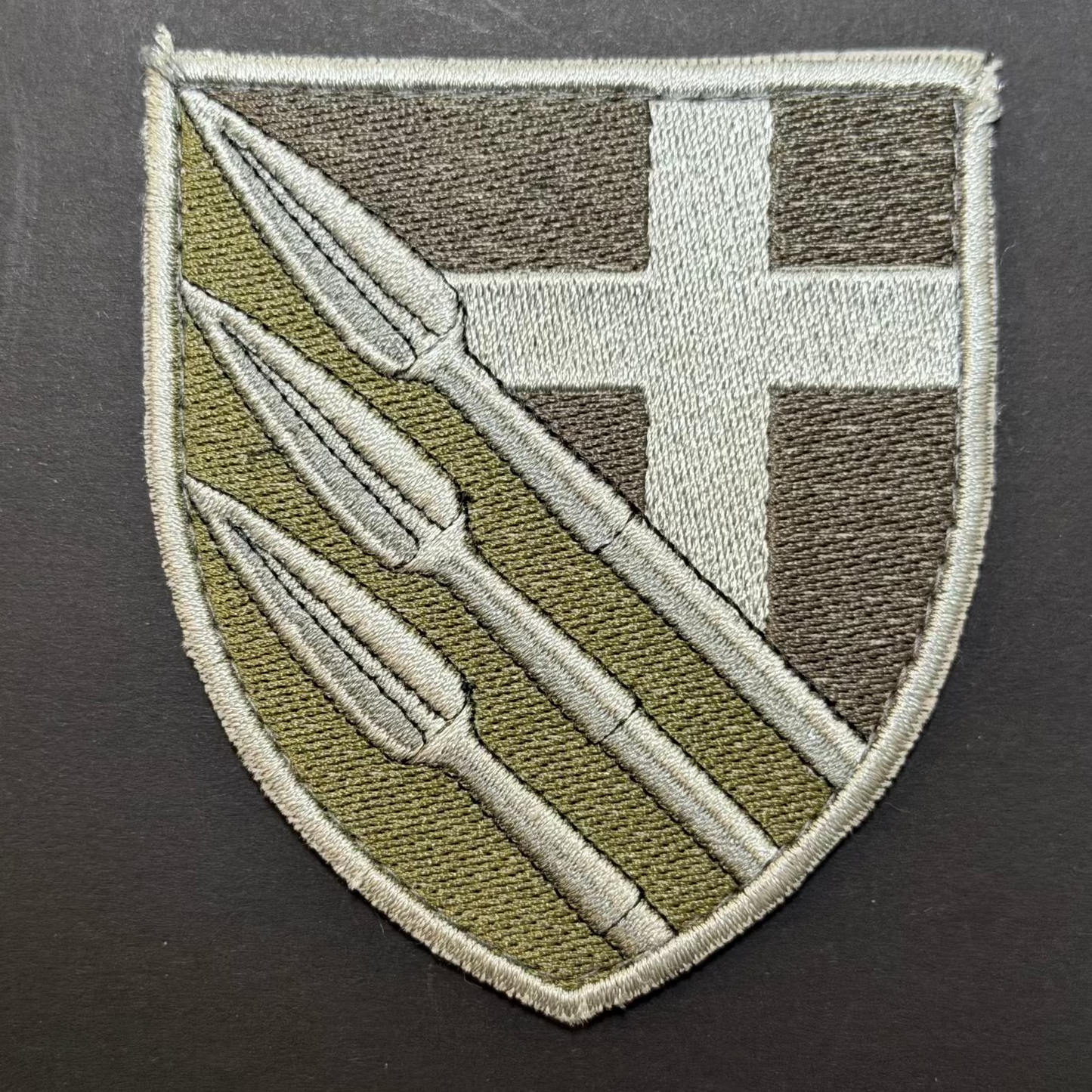 Embroidered patch with a shield design featuring a spear and cross on a dark background