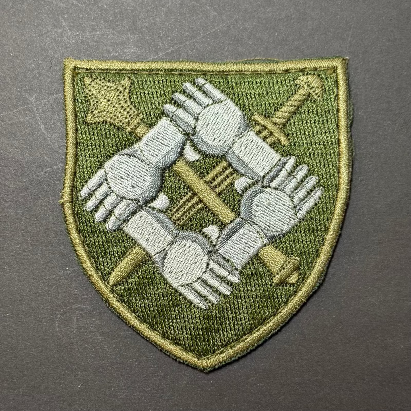 Embroidered patch with interlocking hands and crossed swords on a green shield shape, against a gray background.
