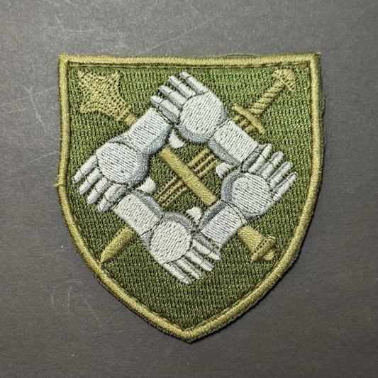 Embroidered patch with interlocking hands and crossed swords on a green shield shape, against a gray background.