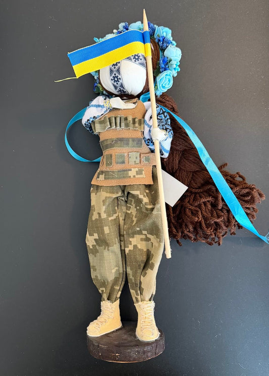 Decorative figure with camouflage outfit, blue and yellow flag, and floral headpiece against a dark background.