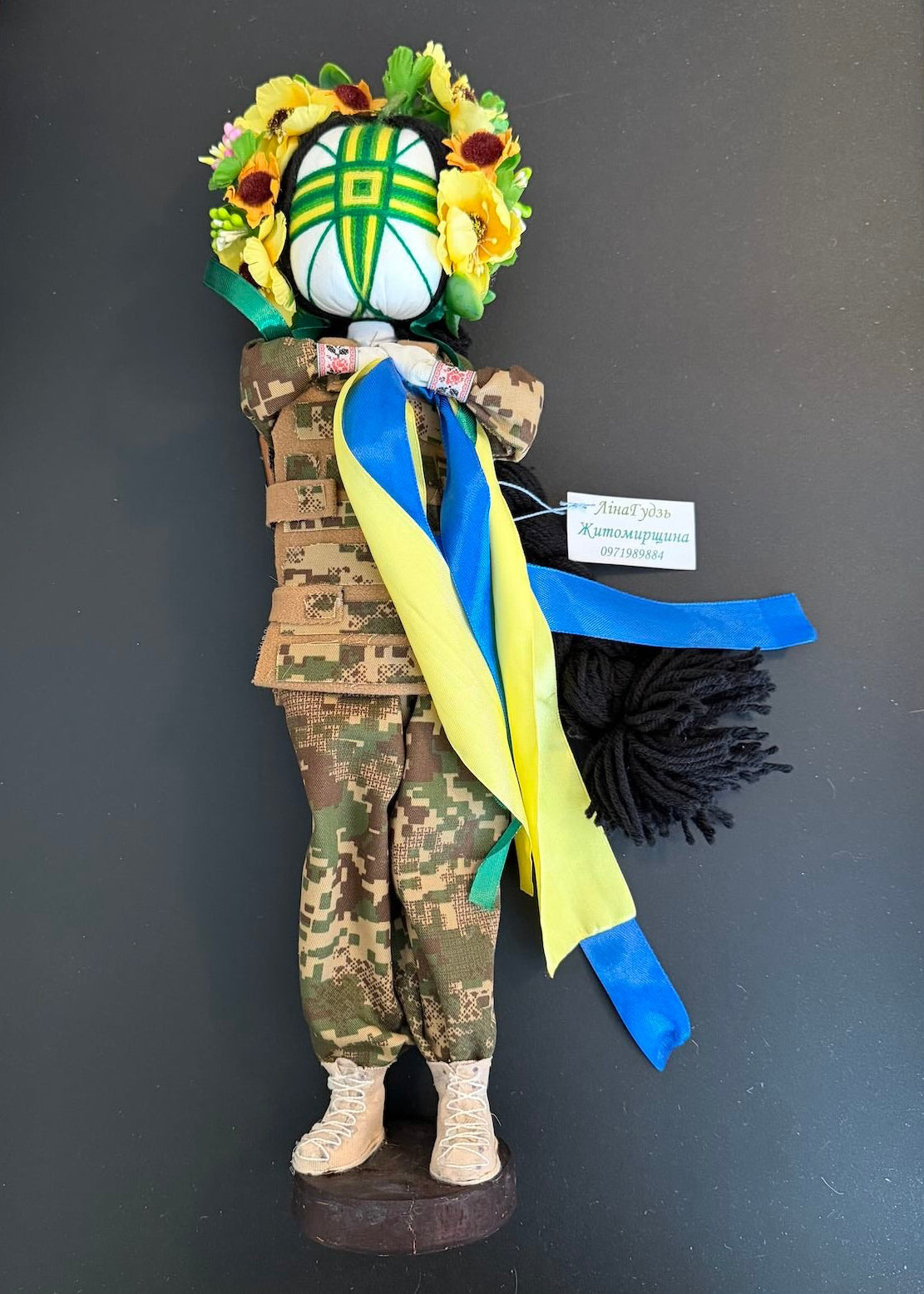 Decorative figure with camouflage attire, flowers, and colorful ribbons on a dark background