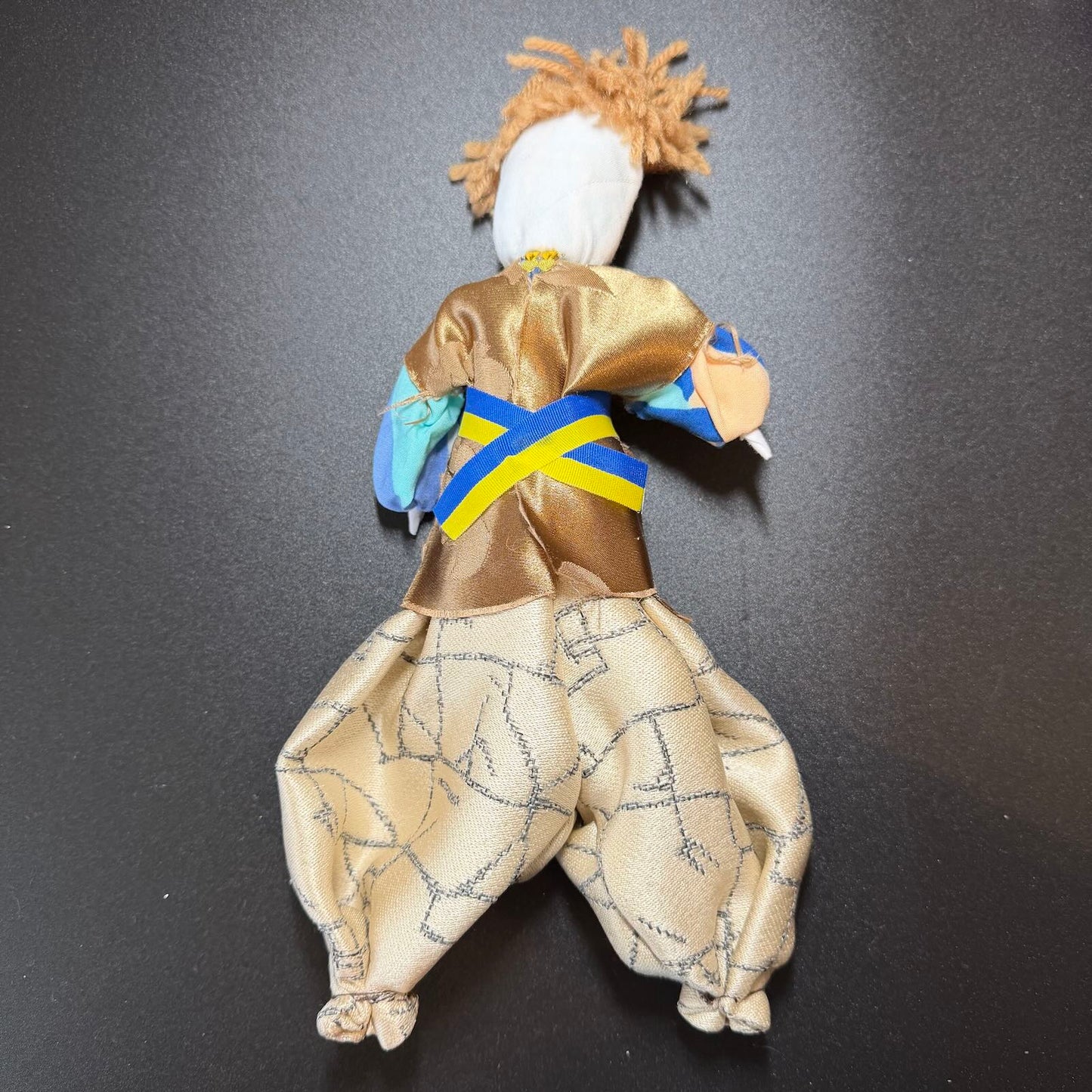 Textile figure with a gold outfit and blue sash on a black background