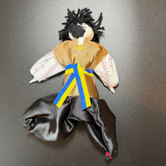 Handmade doll with black hair, brown outfit, and colorful sash on a dark background