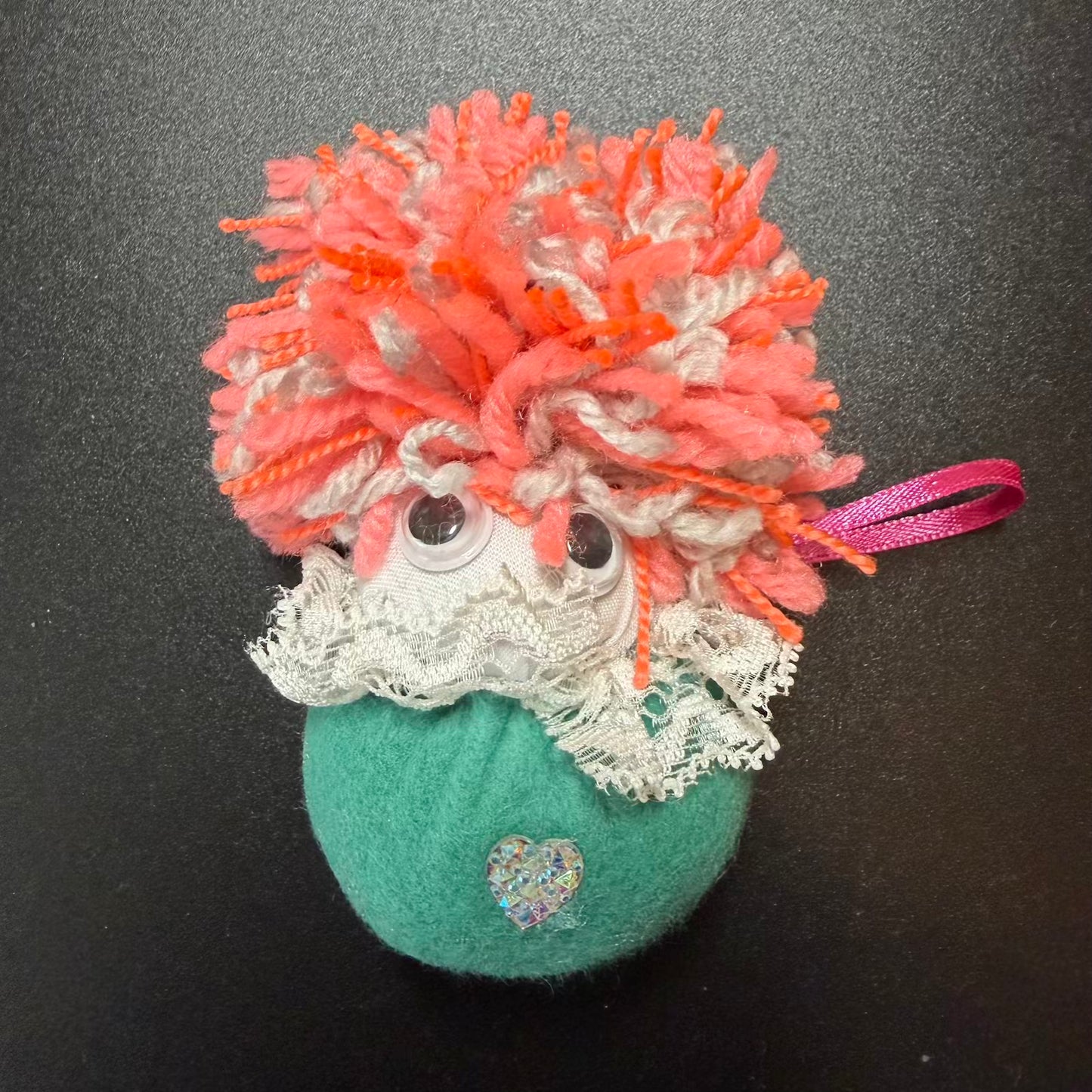 Decorative ornament with pink and orange hair, green body, and heart-shaped charm on a black background