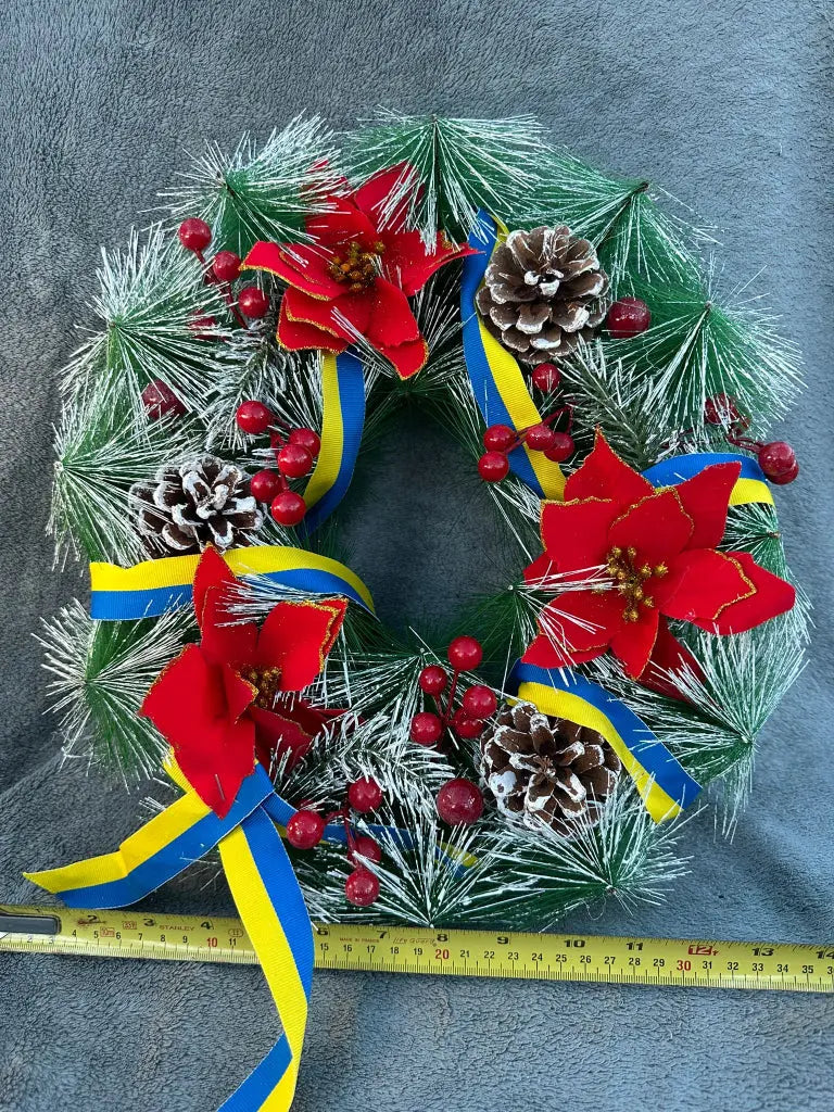A handcrafted Christmas wreath with green pine branches, red and white poinsettias, and blue and yellow ribbons, against a grey background.