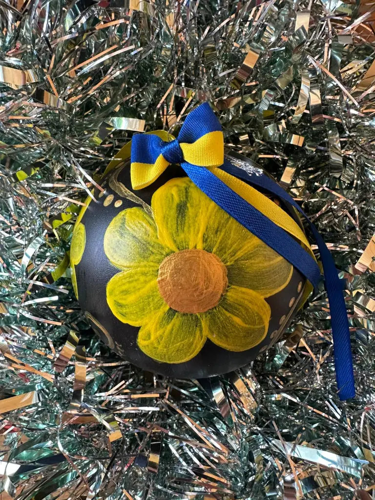 A hand painted Christmas bauble with a flower design, yellow and blue accents, and a blue bow on top, set against a backdrop of glittery decoration.