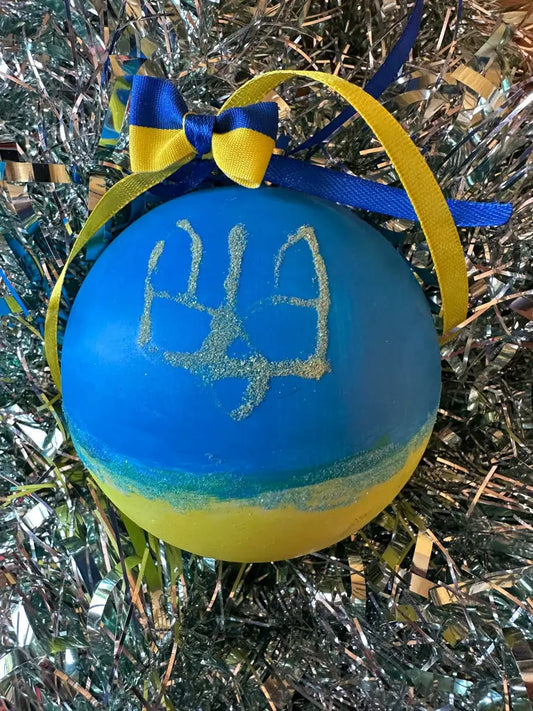 A hand painted Christmas bauble with a blue background and yellow and gold details, featuring a stylized face and a bow with blue and yellow ribbons.