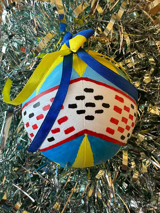 A hand painted Christmas bauble with a geometric pattern featuring blue, yellow, and red colors, adorned with a blue ribbon and yellow flowers, set against a glittery background.